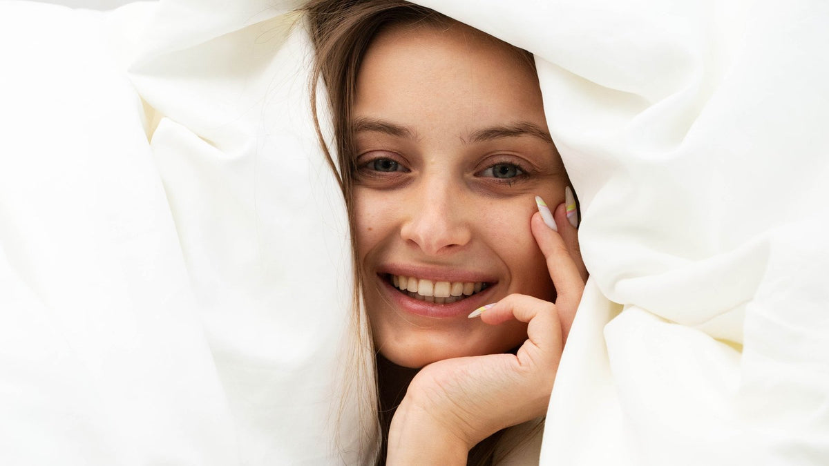 Smiling woman snuggled under a soft white cloud comforter
