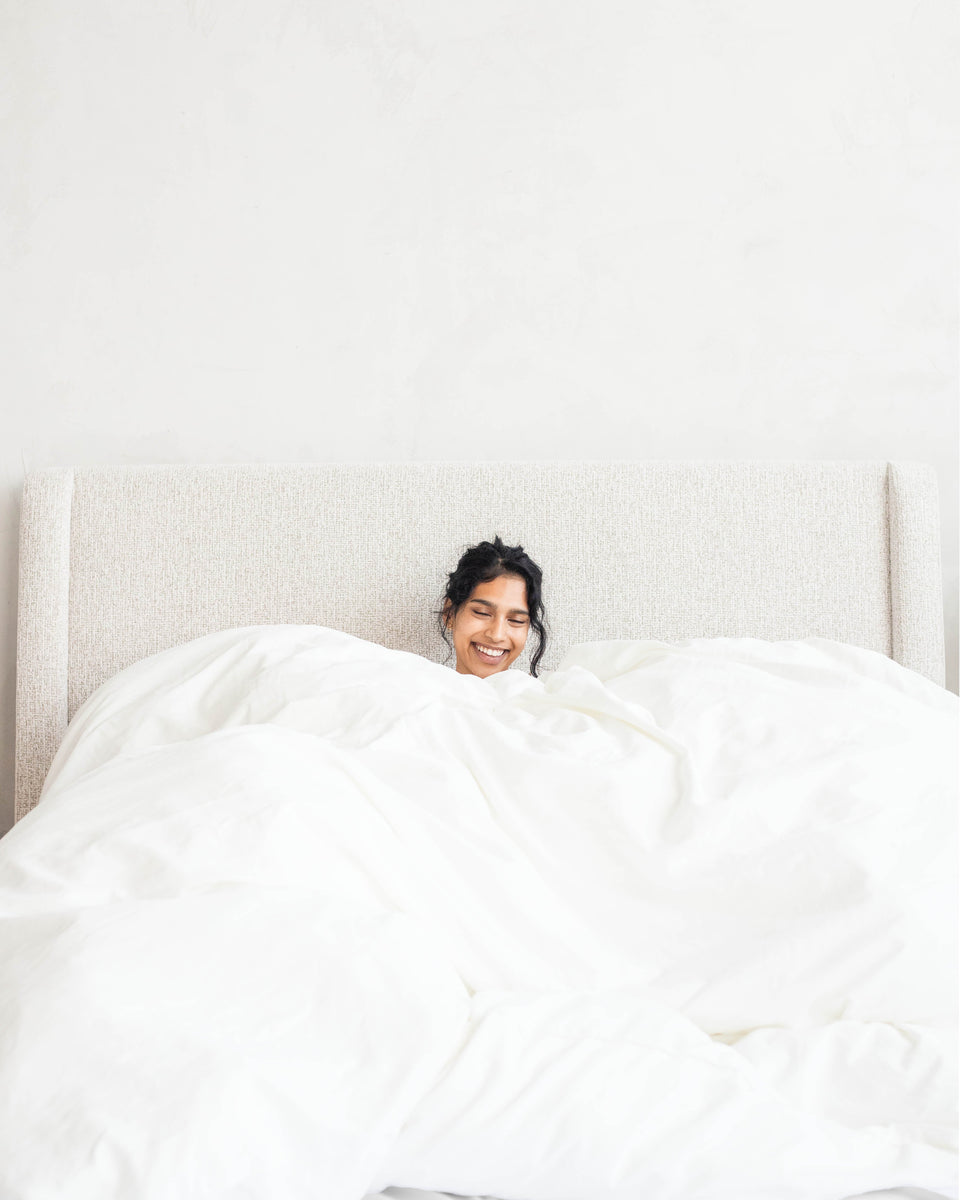 A smiling woman peeks out from under a full, billowy white duvet on an upholstered bed