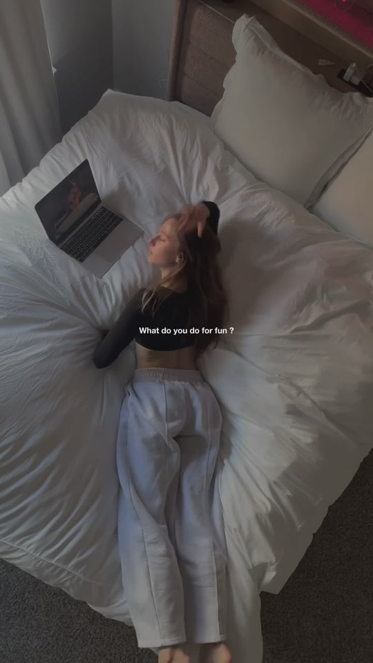 A woman stretches out on a fluffy white duvet with a laptop, then curls up under it