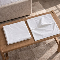 Two empty white cotton percale Cloud Pillowcases folded flat on a wooden bench, one with its envelope closure turned back